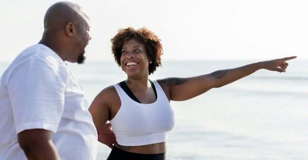 A man and a woman standing by the ocean, with the woman smiling and pointing into the distance.
