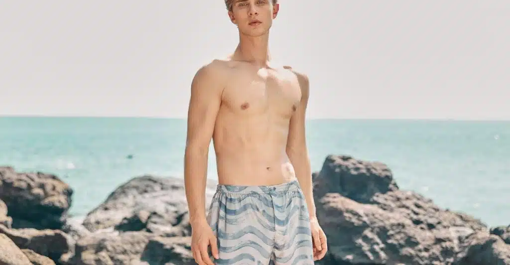A man with a lean build is standing on a rocky beach, wearing light blue and white patterned boxer-style shorts.