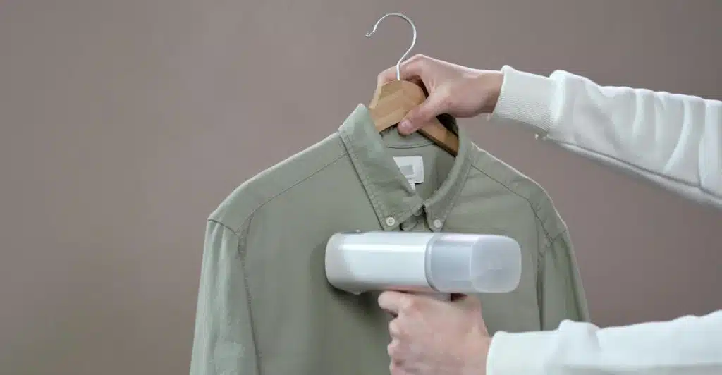 A person holding a steamer to a green collared shirt on a hanger.
