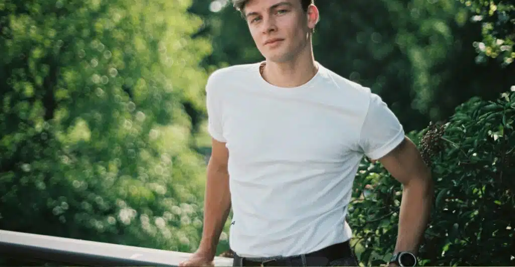 A man in a white t-shirt and dark pants, looking forward with trees in the background.