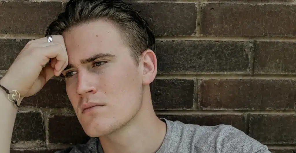 A man wearing a ring and a bracelet, leaning his head on his hand against a brick wall.