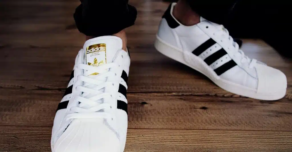 A pair of white Adidas sneakers with black stripes on a wooden floor.