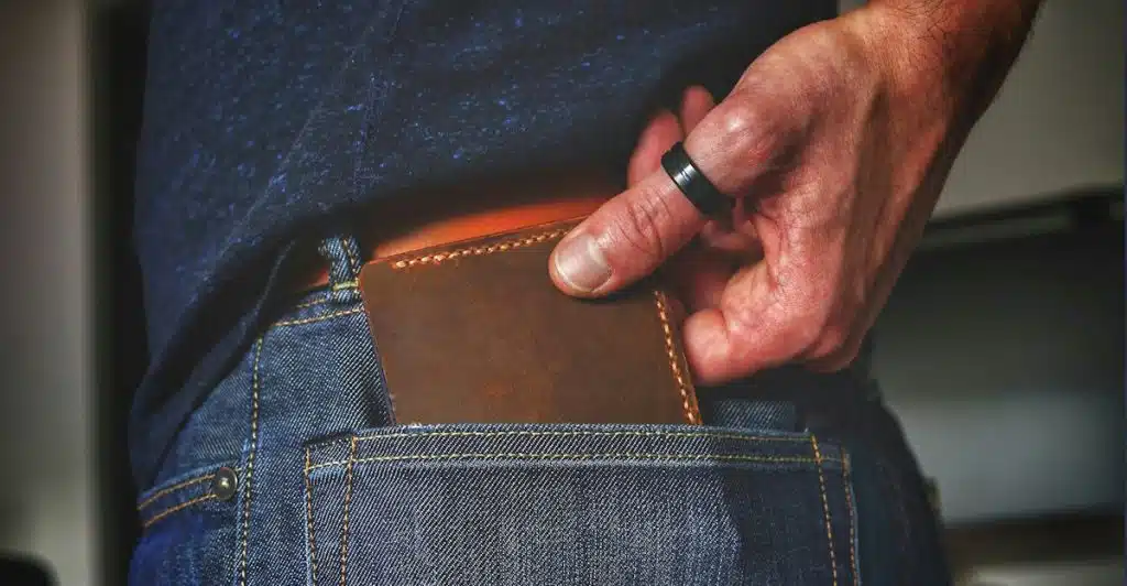 A hand pulling a brown leather wallet with stitching from the back pocket of a pair of blue jeans.