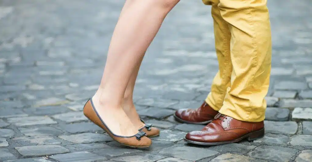 The lower half of two people standing, one wearing brown ballet flats and the other wearing brown square-toe dress shoes