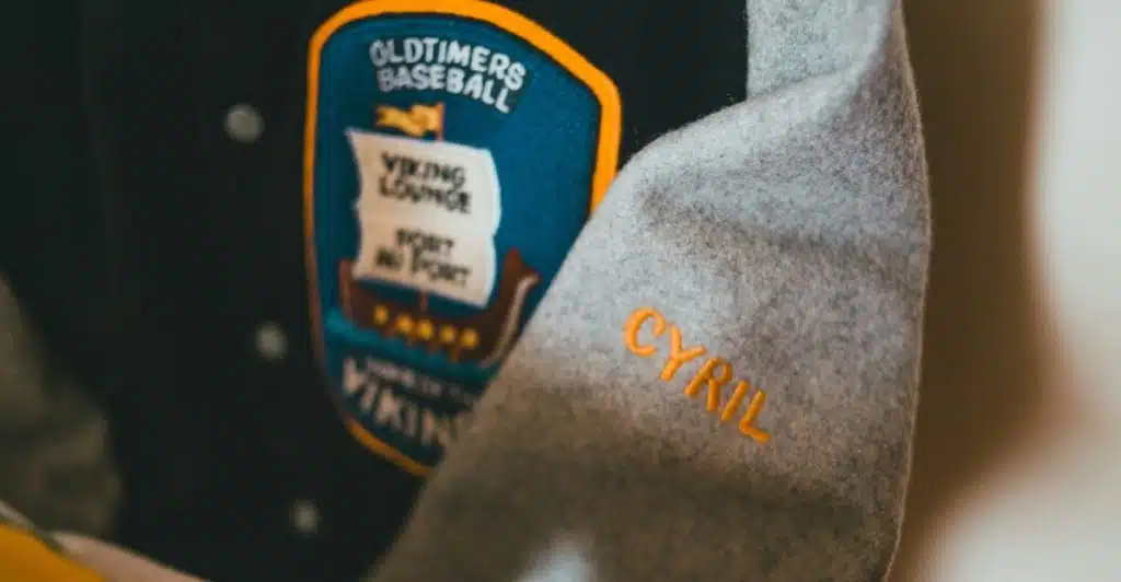 A close-up of the sleeve of a letterman jacket with a "OLDTIMERS BASEBALL" patch and "CYRIL" embroidered on the sleeve.