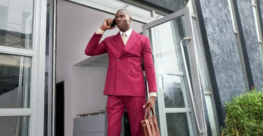 A man in a loose-fitting burgundy suit holding a briefcase and talking on a phone while standing in a doorway.