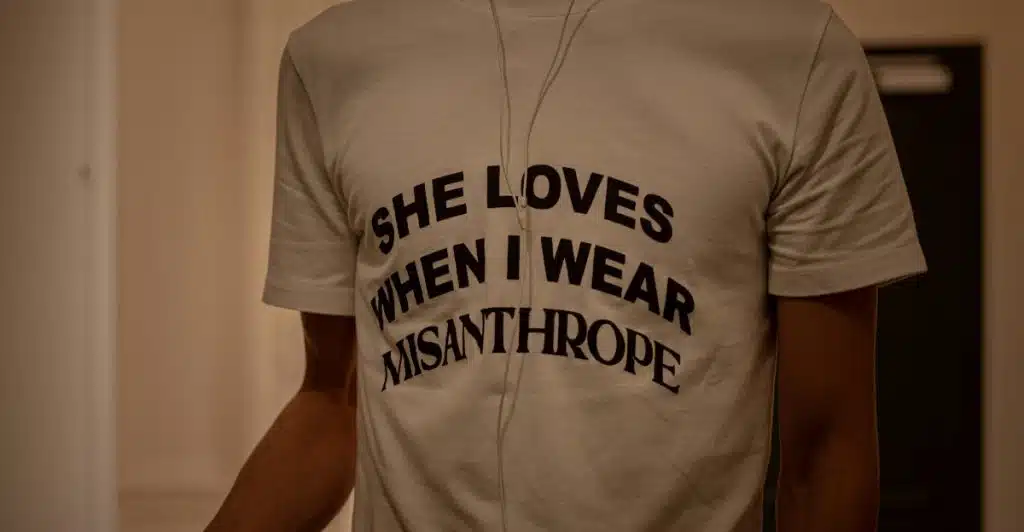 A man wearing a white t-shirt with the words "SHE LOVES WHEN I WEAR MISANTHROPE" printed on it