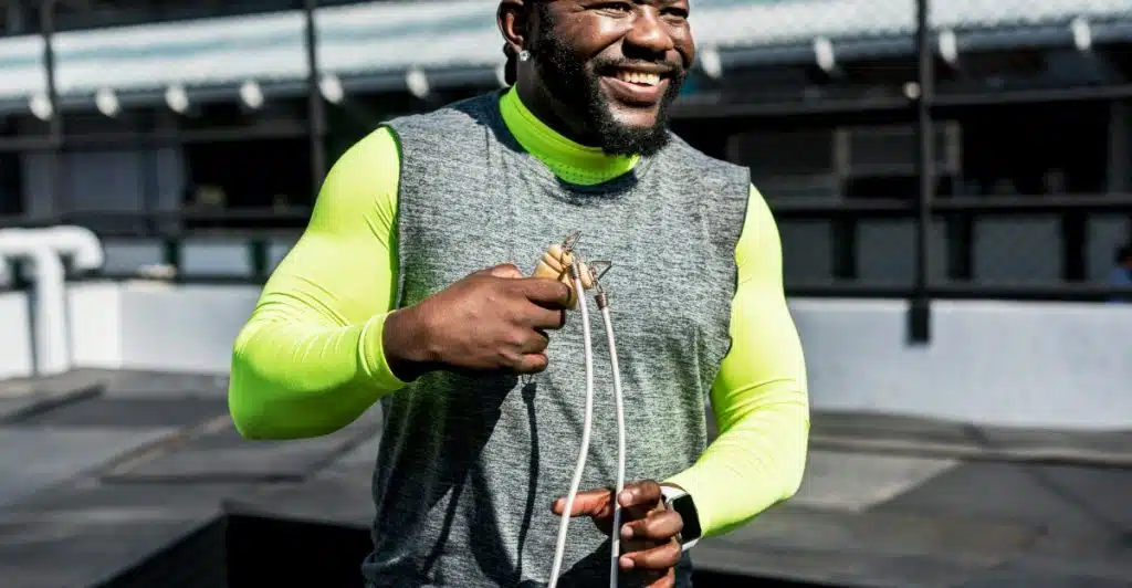 A smiling man in a grey sleeveless top over a neon green long-sleeved shirt holding a jump rope.