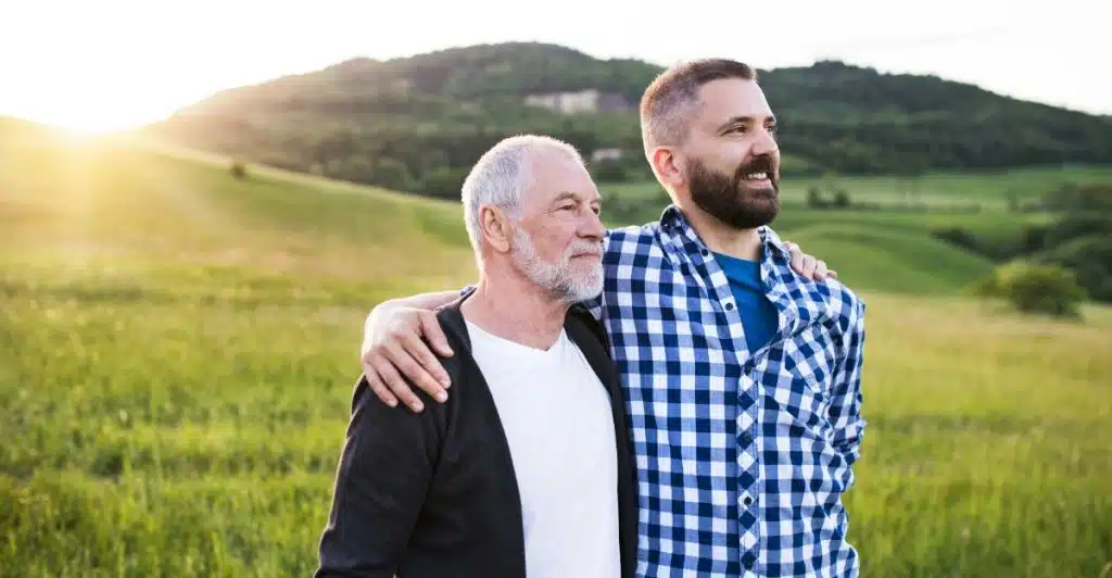 Two men, one older with white hair and a younger man with a beard, standing in a grassy field with a sunset in the background.