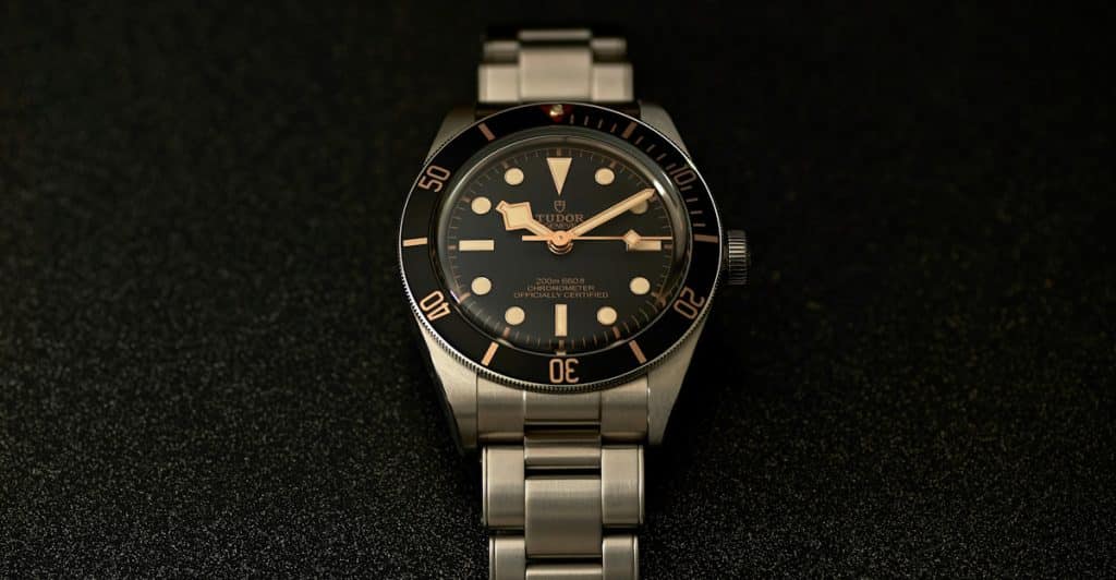 A close-up view of a Tudor Black Bay watch with a black dial and a stainless steel bracelet on a textured dark surface.