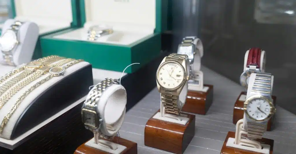 Several luxury watches displayed in individual cases on a shelf, with some watch boxes visible in the background.