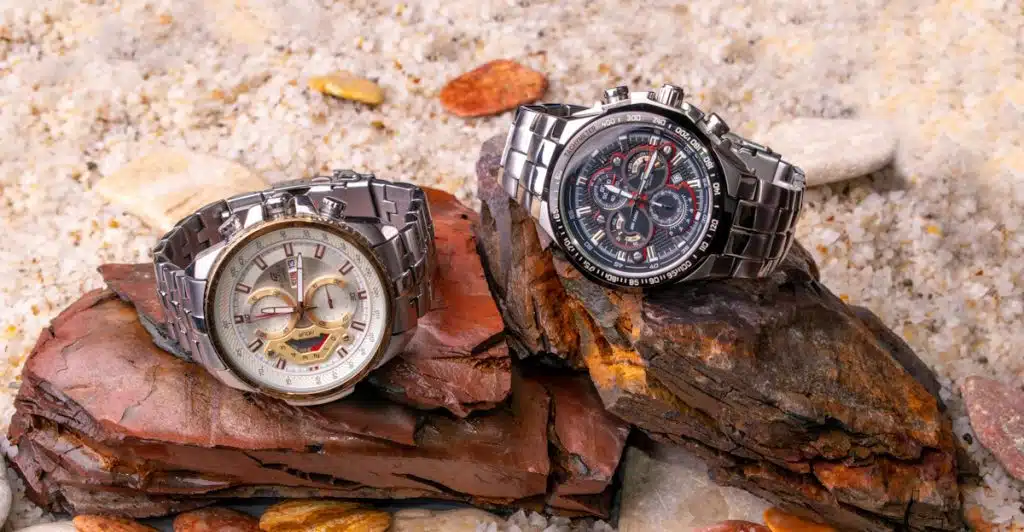 Two luxury watches, one silver and one black, placed on a bed of rocks and sand.