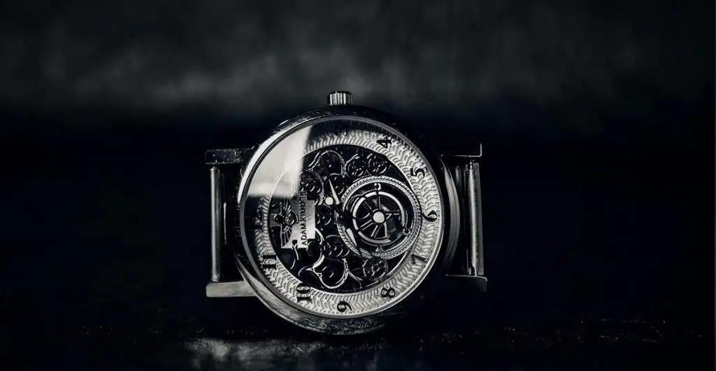 A close-up black and white image of a mechanical wristwatch placed on a dark surface.