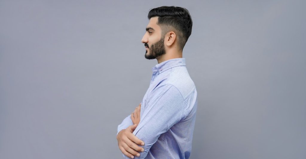 A man with a taper fade haircut and a beard standing sideways with his arms crossed.