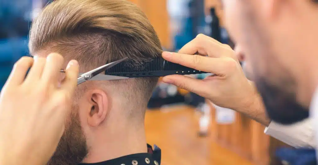 A man getting a brushed back style haircut by a barber using scissors and a comb.