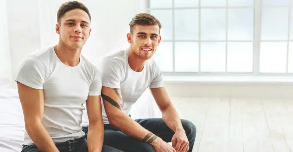 Two men with Ivy League haircuts and matching outfits sitting on a bed and smiling.
