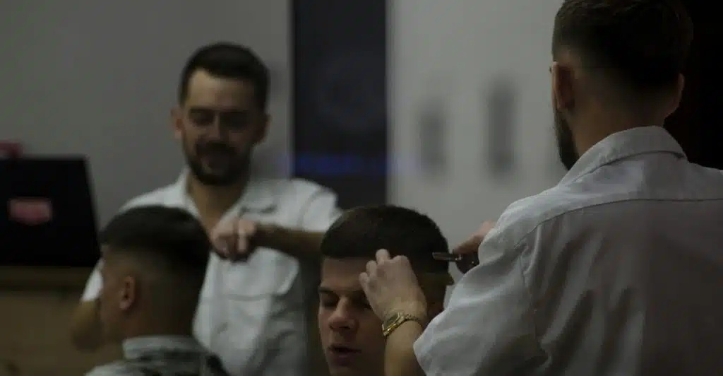 Men getting Caesar cuts at a barbershop, with one barber cutting a man's hair and another man reflected in a mirror.