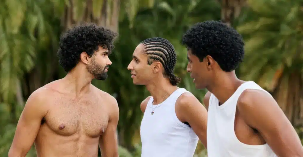 Three men with short afro or close curl fade haircuts, one shirtless and two in white tank tops, standing outdoors.