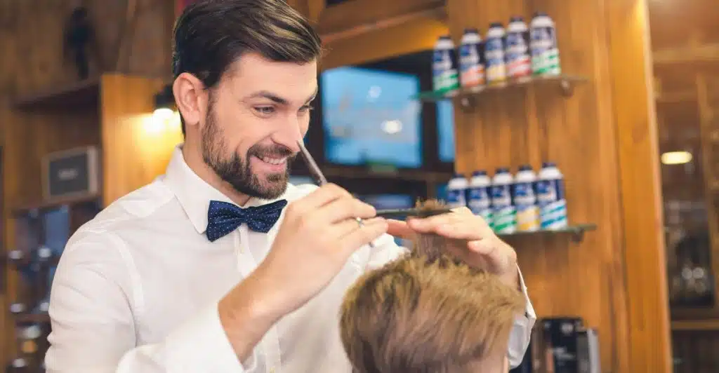 A smiling barber wearing a bow tie cutting a man's hair with scissors in a barbershop.