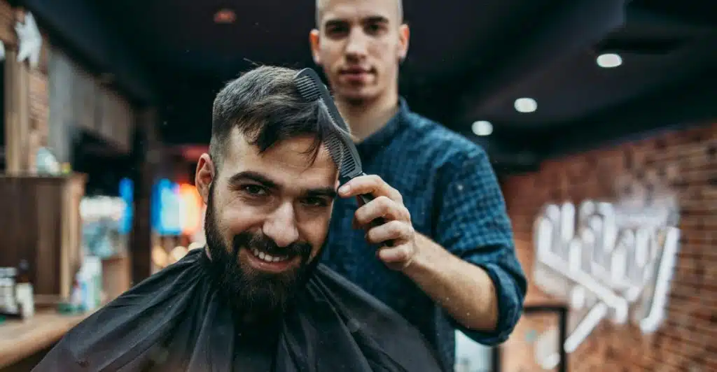 A smiling man in a barber's chair with a barber combing his beard.