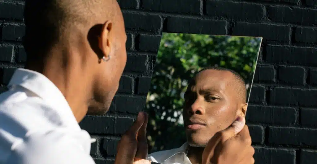 A man with a buzz cut looking at his reflection in a mirror against a black brick wall.