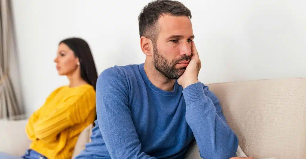 A man and a woman sitting apart on a sofa, with their backs to each other, suggesting a disagreement or emotional distance between them.
