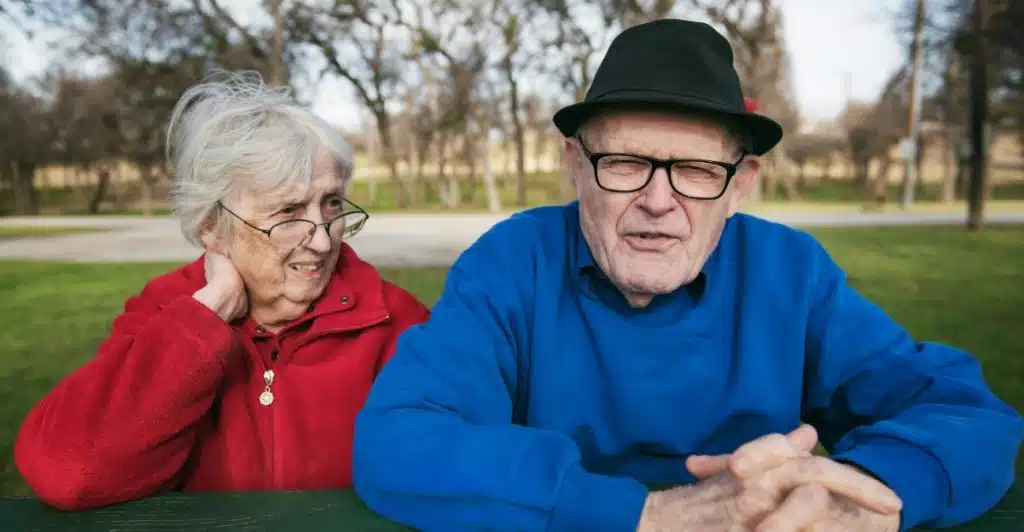 An elderly couple seated at an outdoor picnic table, with the woman looking towards the man, who is looking towards the camera.