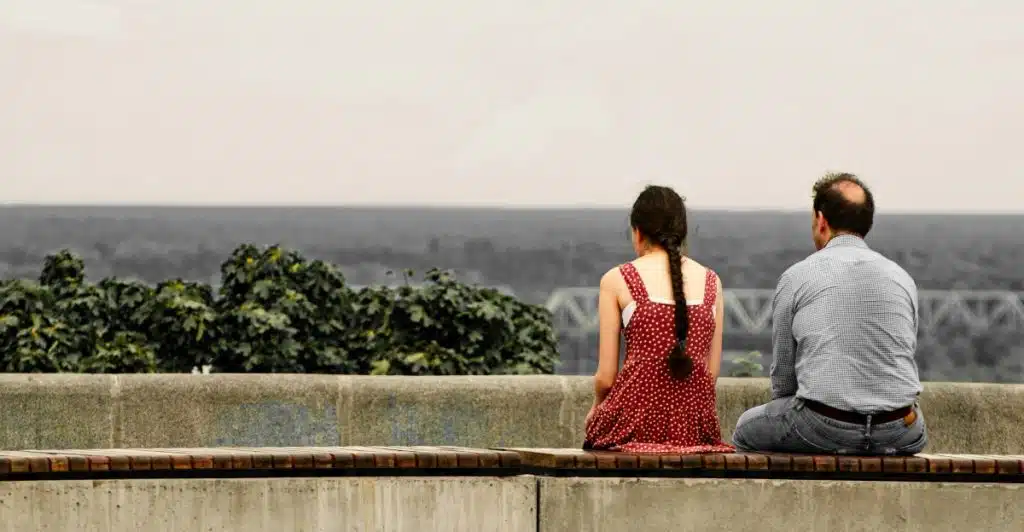 A man and a woman seated side by side on a bench, with their backs to the viewer, gazing out at a vast, open body of water under a cloudy sky.