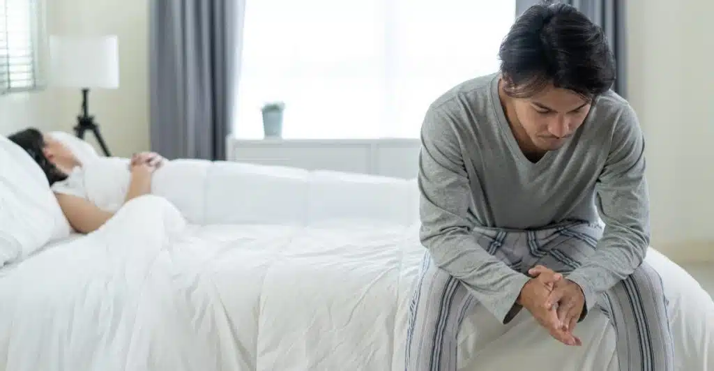 A man is sitting on the edge of a bed, looking contemplative, while a woman is lying in the background, appearing to be asleep or resting.
