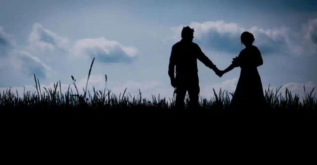A silhouetted couple holding hands standing in a field of tall grass against a dramatic sky with clouds.