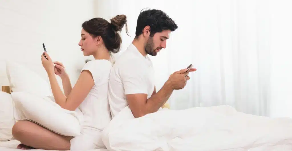 A man and a woman sitting on a bed with their backs to each other, both engrossed in their mobile phones.
