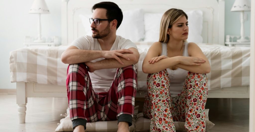 A man and a woman sitting on the floor with their backs against a bed, looking away from each other, suggesting a disconnect.