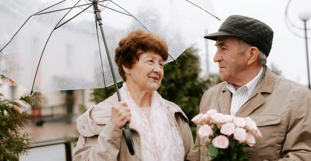 An elderly couple standing together under a clear umbrella, with the man holding a bouquet of pink roses, both looking at each other.
