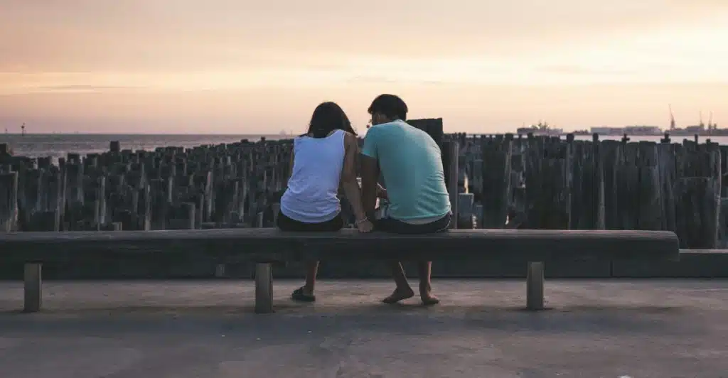 A couple sitting on a wooden bench by the water, looking out at the sunset over a distant pier.
