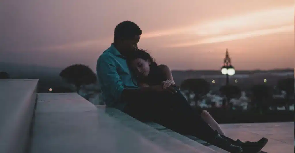 A couple embracing outdoors at dusk, with the man holding the woman close as she rests her head on his shoulder.