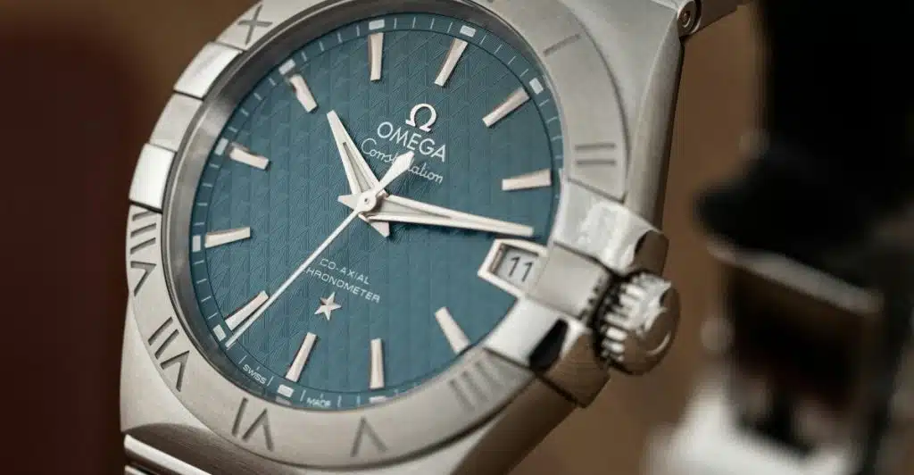 A close-up of an Omega De Ville Prestige watch with a textured blue dial, silver case, and Roman numerals, angled to show the watch face and part of the bracelet.