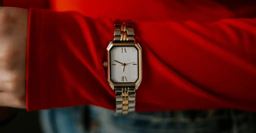 A wrist wearing a rectangular Cartier Tank Must Small watch with a two-tone bracelet and a white dial with Roman numerals, set against a red sleeve and blue jeans.