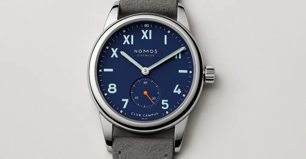 A Nomos Club Campus 36mm watch with a deep blue dial, mixed Arabic and Roman numerals, and a grey suede strap, presented on a light background.