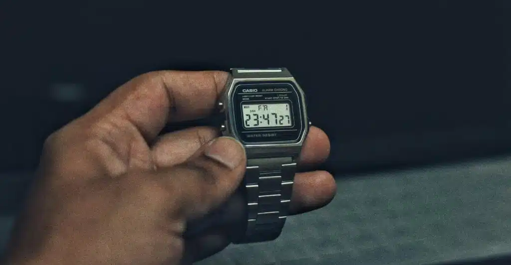 A person's hand holding a silver Casio A168WA digital watch, displaying the time and date against a dark, blurred background.