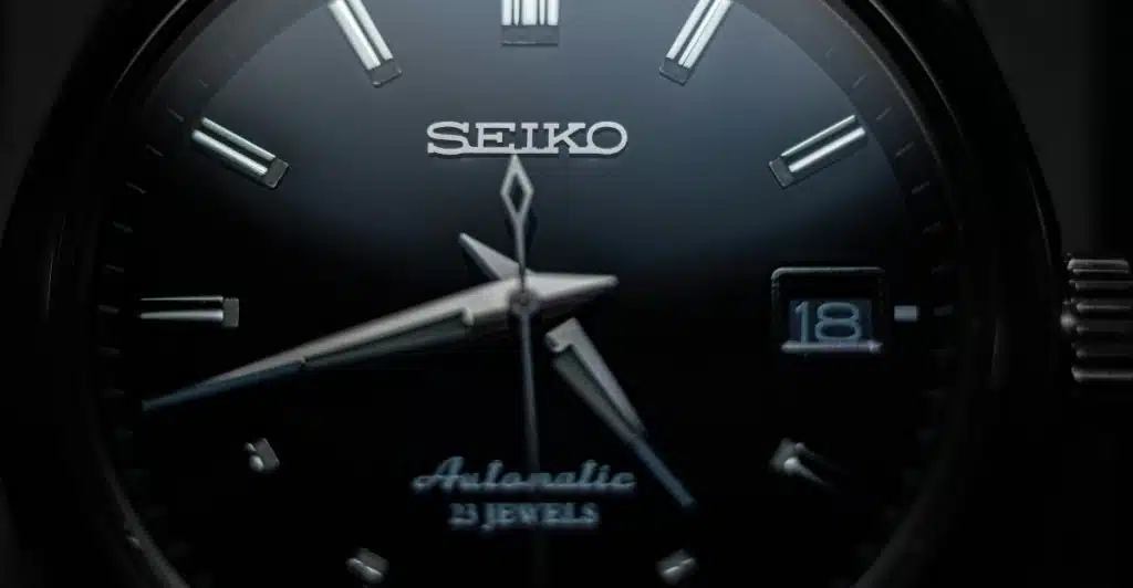 A close-up of a Grand Seiko SBGX261 watch face, featuring a black dial, polished silver hour markers, and a date window, with visible text indicating "SEIKO" and "Automatic".