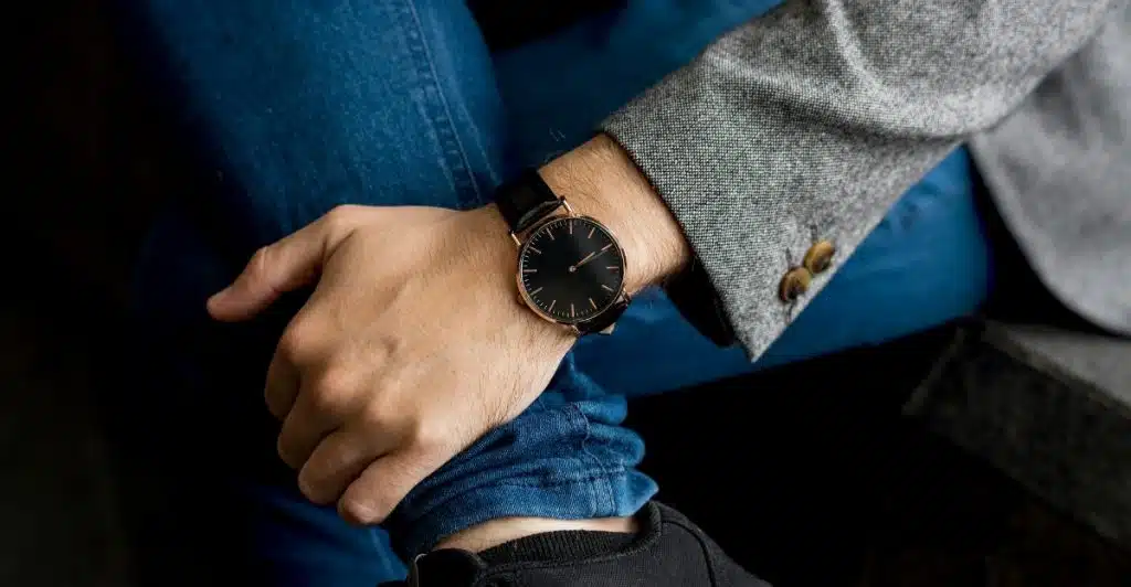 A man wearing a Longines DolceVita watch with a black strap and a gold-tone rectangular case and black dial, seen from a seated position with blue jeans and a grey blazer.