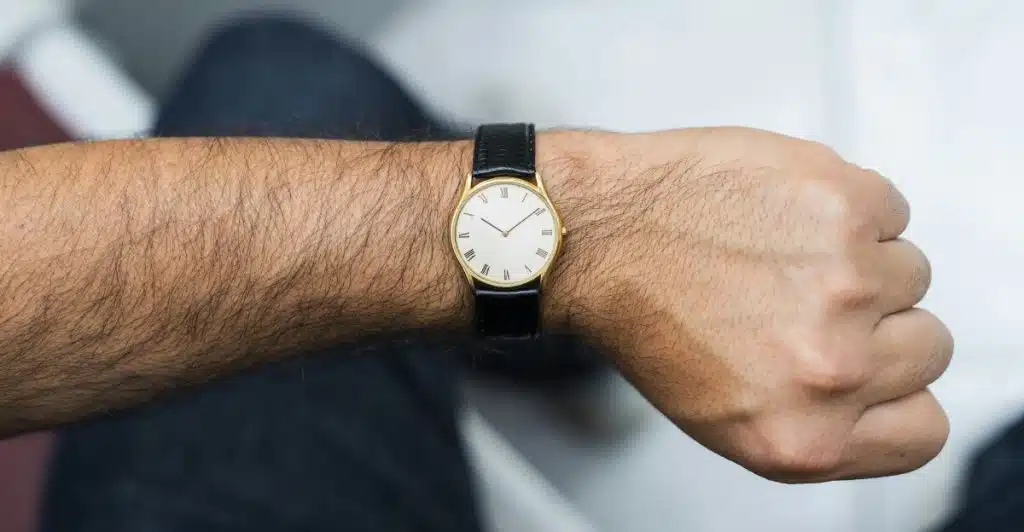 A man's forearm with a gold-tone watch with a white dial and black leather strap, positioned to show the watch clearly on the wrist.