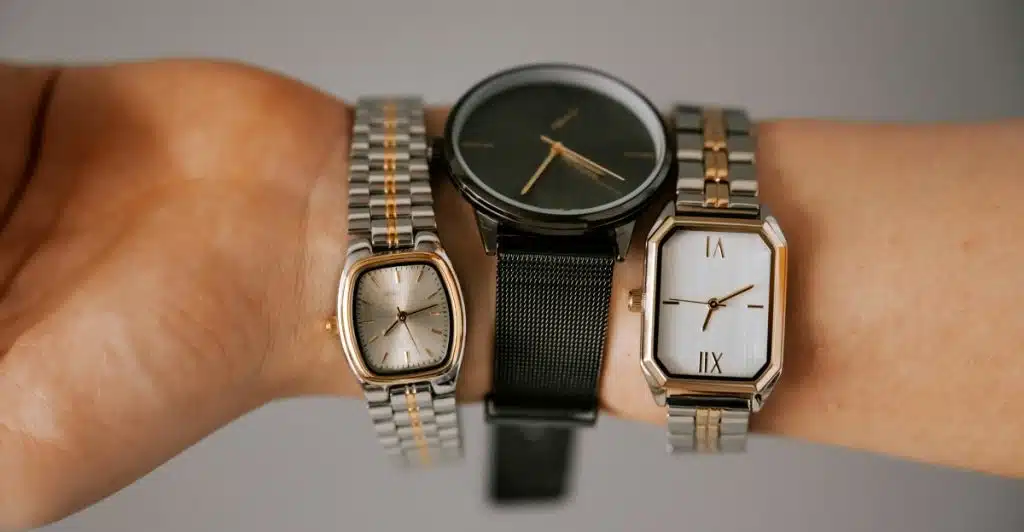 Three watches displayed side-by-side on a wrist: a rectangular gold and silver watch, a round black watch with a mesh strap, and another rectangular silver and gold watch with Roman numerals.