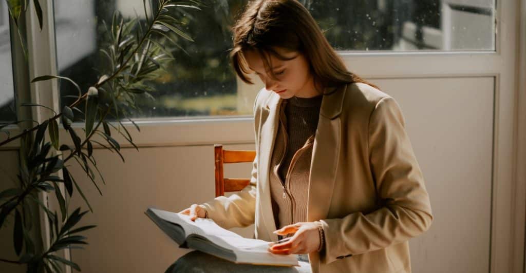 A young woman sitting indoors, holding and reading a physical book.