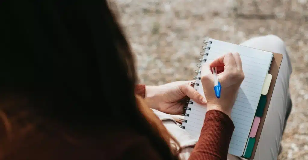 A person from the shoulders down, writing in a notebook with a pen while sitting outdoors.