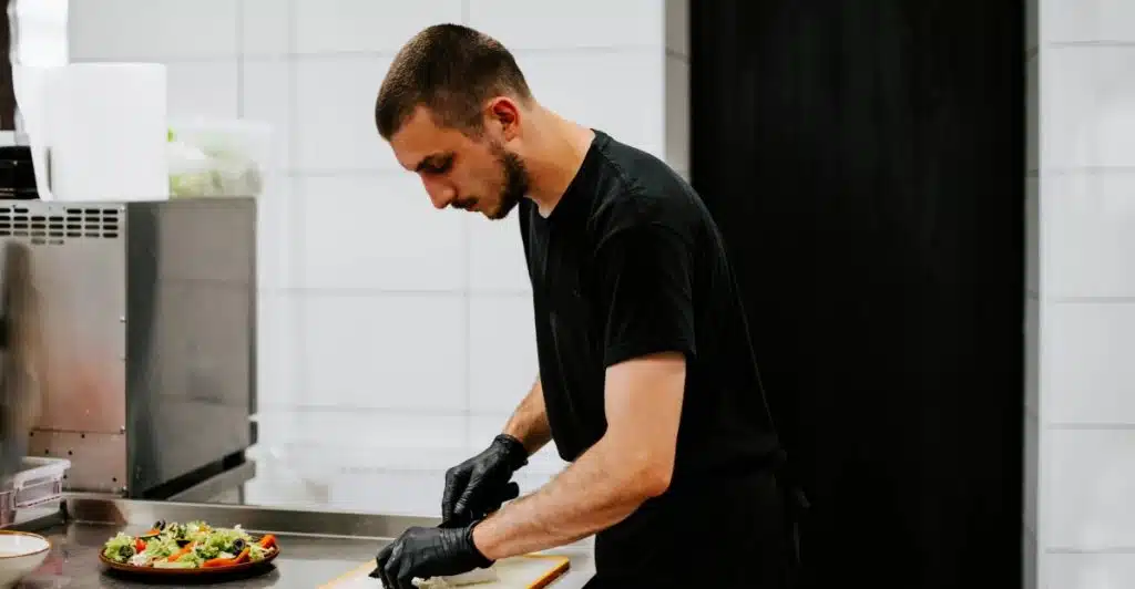 A man in a black t-shirt and black gloves is chopping food on a counter in a kitchen.