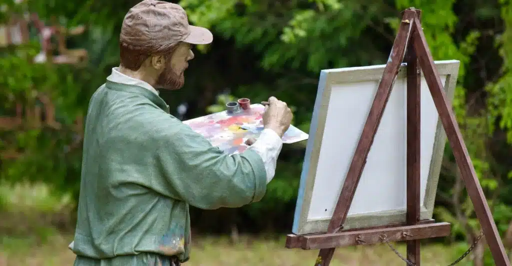A man wearing a hat and green shirt painting on an easel in a natural outdoor setting.