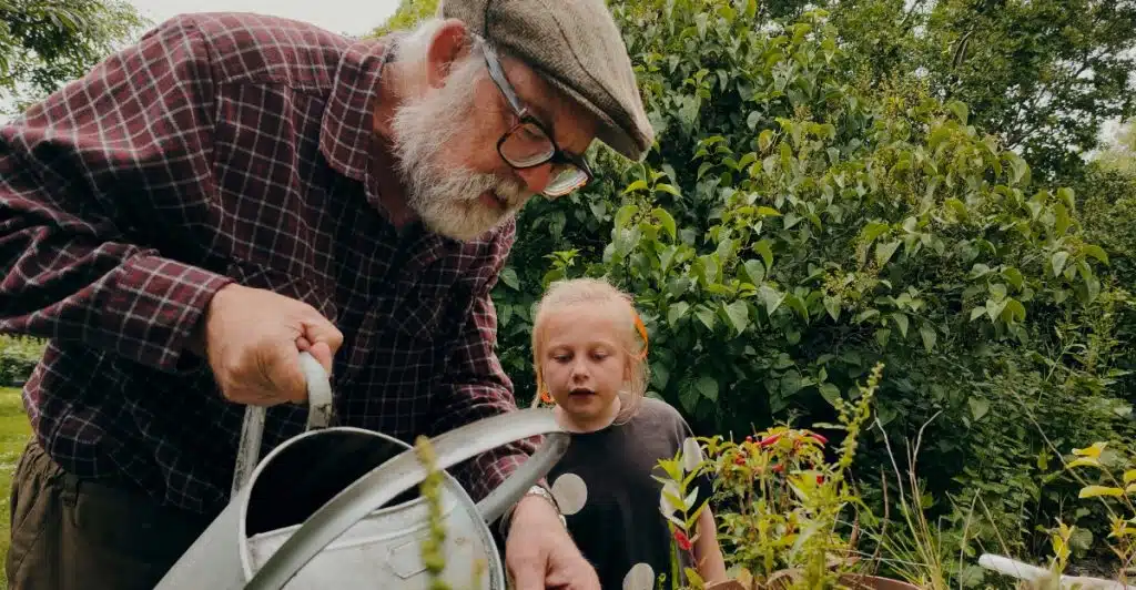 An older man with a beard and a young child together in a garden, with the man holding a watering can.