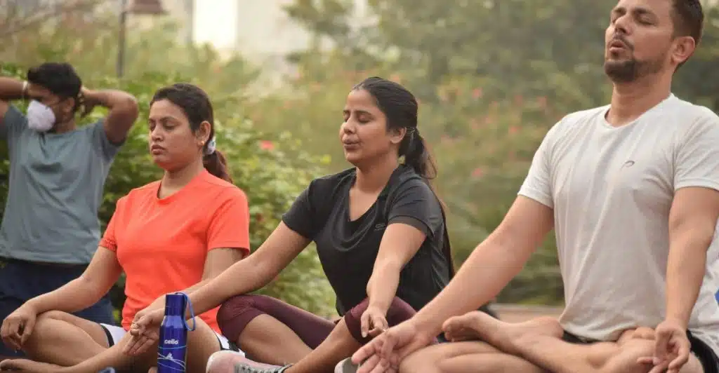 Three individuals sitting in a meditation pose on yoga mats outdoors, with one person in the background stretching.