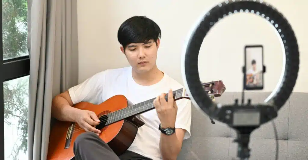 A young man sitting on a couch, playing an acoustic guitar, with a ring light and a phone visible in the foreground.